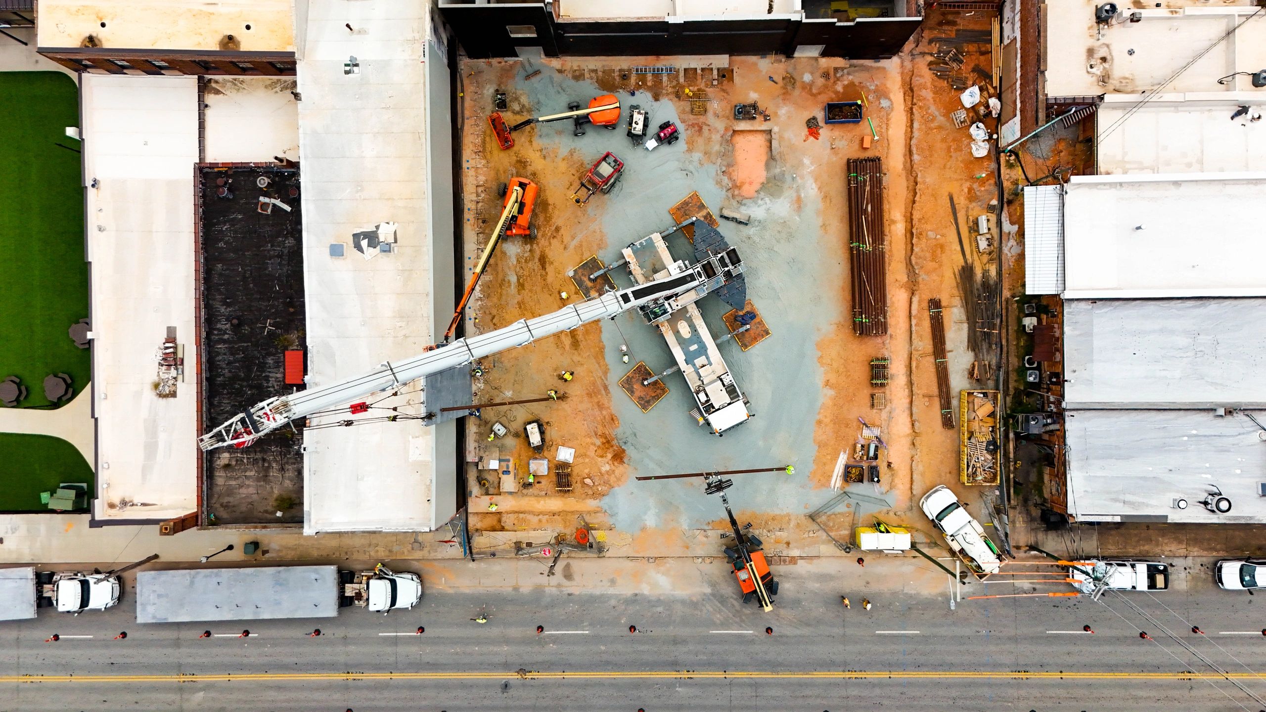 Straight-down view of a crane, workers, lifts, and staged materials on a construction site between existing buildings.